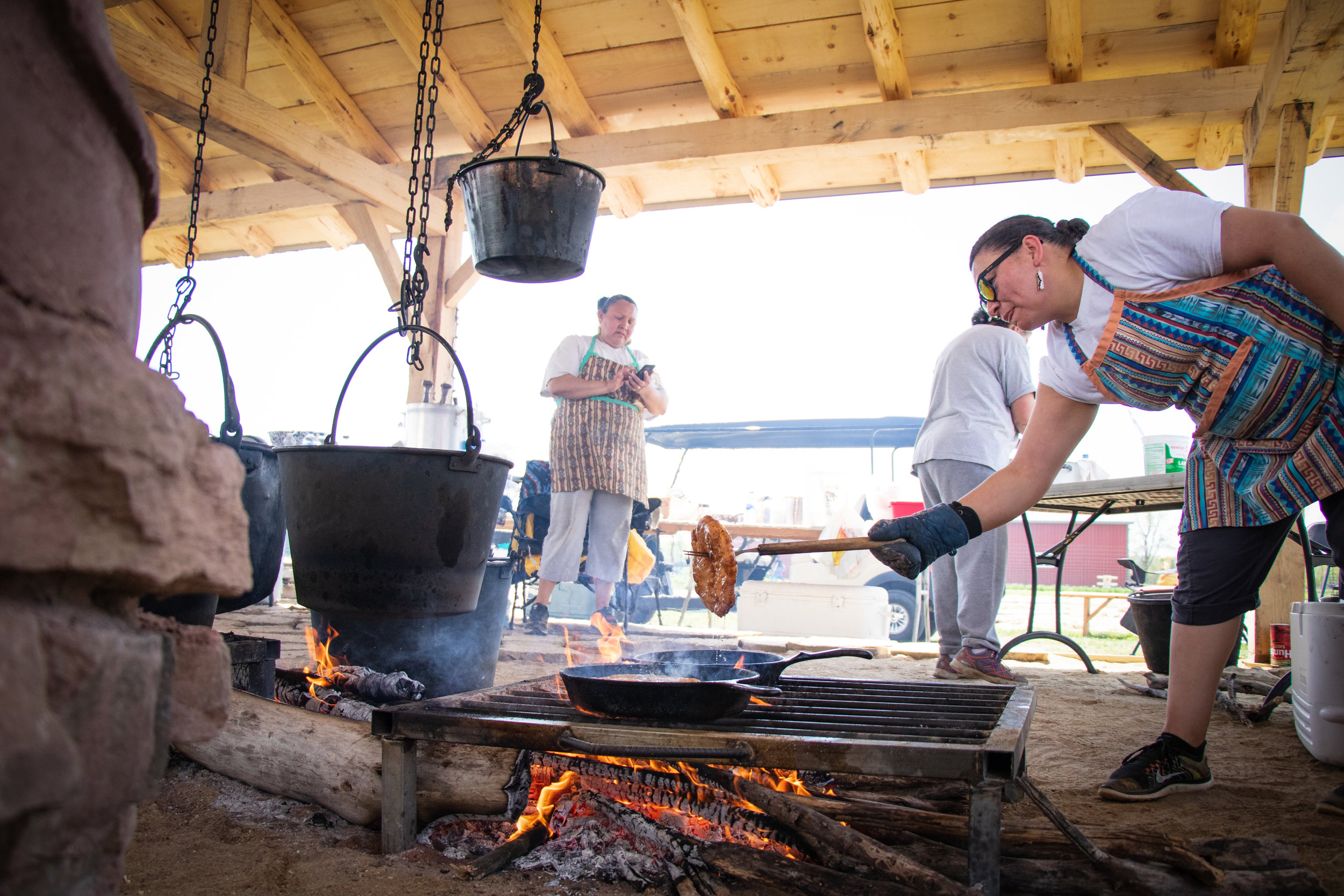 Cooking demonstration at an outdoor kitchen, featuring a woman in an apron stirring food over an open fire, with black pots hanging above. Two other individuals are visible in the background, engaging in food preparation. The setting showcases traditional cooking methods in a rustic environment.