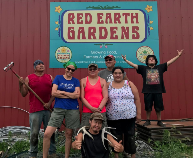 Group of six individuals standing in front of the Red Earth Gardens sign, promoting sustainable farming and community engagement. The team is wearing casual clothing and smiles, showcasing their commitment to growing food and supporting the Meskwaki community.